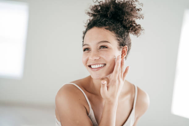 Happy young woman applying facial cream in a brightly lit room