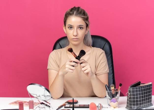 Beauty blogger comparing makeup brushes at her workspace with various cosmetics laid out on a table, against a pink background