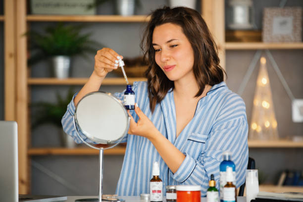 A woman using a dropper and bottle of serum while sitting at a desk with a mirror and various beauty products.